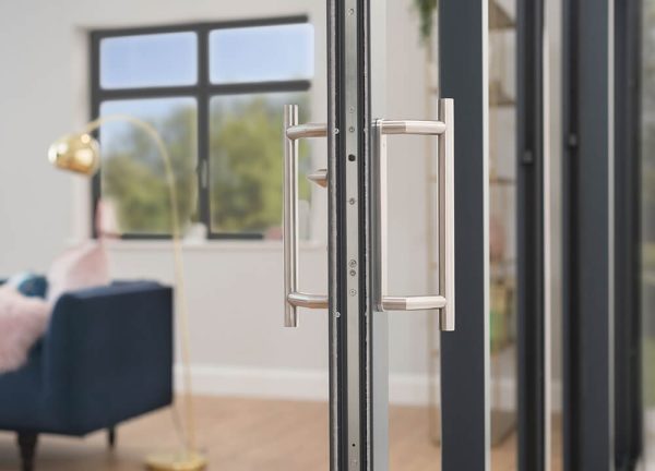 Close-up of a bifold door handle in a satin nickel finish, with a navy sofa and brass floor lamp visible through the glass.